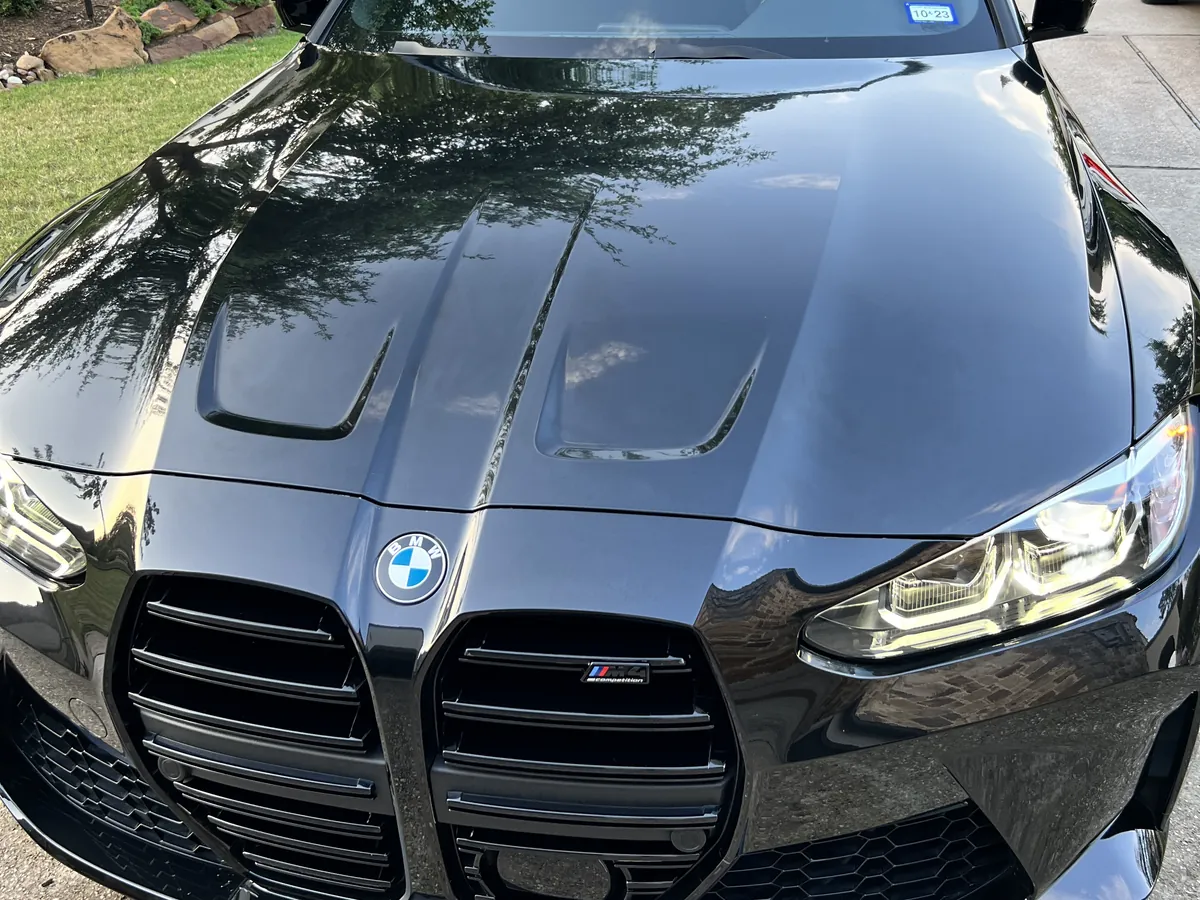Black BMW M4 convertible after an Azul detail, its glossy hood reflecting the trees in a driveway in The Woodlands