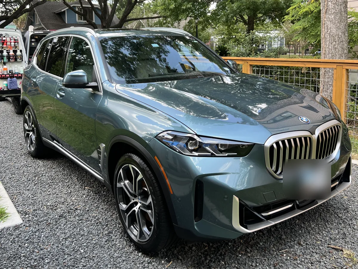 Blue-gray BMW X5 after an Azul detail, on a gravel driveway under oak trees in the Houston Heights, with the Azul van behind it