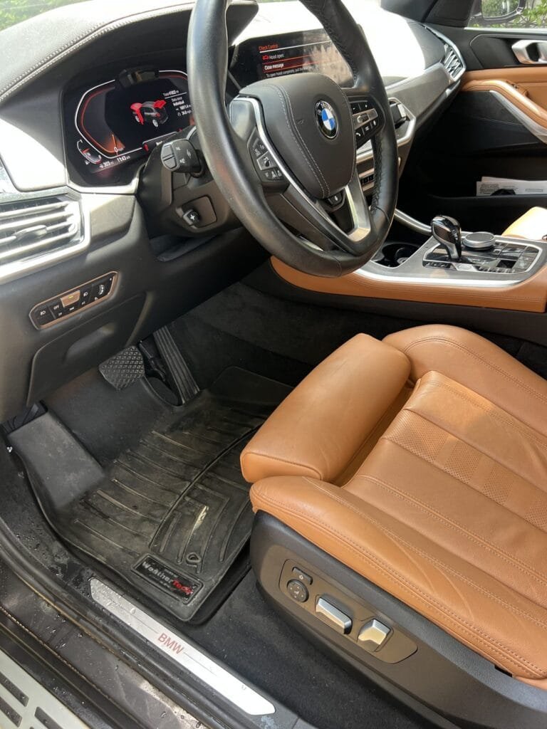 BMW X5 driver's side before interior cleaning: dirty floor mat and dusty trim