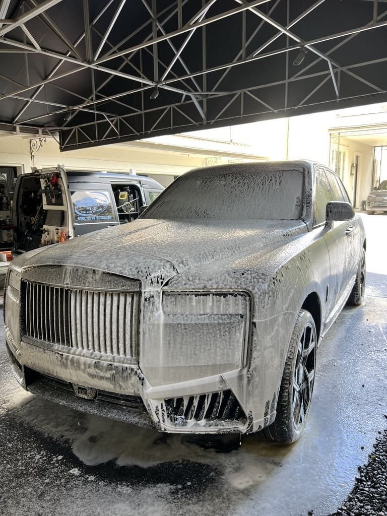 Rolls-Royce Cullinan covered in wash foam under a covered driveway, with the open Azul van and equipment beside it